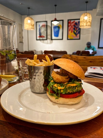 A plated meal featuring a burger with a variety of toppings next to a metal cup filled with French fries. The setting appears to be a cozy dining area with wooden tables and artistic framed prints on the walls, illuminated by hanging lights. A glass pitcher is placed nearby, containing a drink with mint leaves and lemon slices.