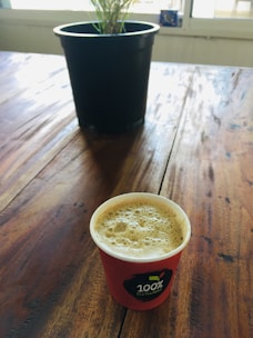 A wooden table with a small plant in a black pot in the background and a red coffee cup labeled '100% Recyclable' in the foreground. The coffee cup is filled with frothy coffee.