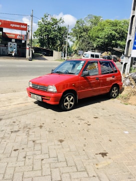 A small red car is parked on a brick-paved area near a road. The car has a license plate reading '302-6413' and black, five-spoke wheels. In the background, there is a service station with signs for 'xpress lube' and 'Havoline', along with trees and vegetation visible in the vicinity.