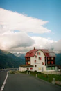 A hotel named 'Galenstock' located in a mountainous area. The building has a rustic appearance with red shutters and a red roof. It's situated next to a winding road that leads into the distance through the mountains. The sky above is partly cloudy, creating a dramatic backdrop to the scene.