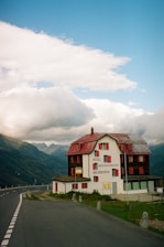 A hotel named 'Galenstock' located in a mountainous area. The building has a rustic appearance with red shutters and a red roof. It's situated next to a winding road that leads into the distance through the mountains. The sky above is partly cloudy, creating a dramatic backdrop to the scene.