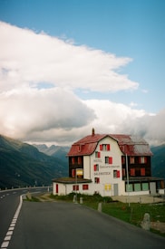 A hotel named 'Galenstock' located in a mountainous area. The building has a rustic appearance with red shutters and a red roof. It's situated next to a winding road that leads into the distance through the mountains. The sky above is partly cloudy, creating a dramatic backdrop to the scene.
