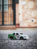 A toy car with green and white coloring, displaying the words 'fast' and '28' on its side, positioned on a textured, gravelly surface. The background is blurred, showcasing a building with a prominent door and brickwork, giving the scene a sense of depth.