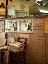 The scene depicts the interior of a pizzeria with a tiled wall and stacks of pizza boxes on chairs. Through a window, a person is seen preparing food in a kitchen. Various posters and drawings are attached to the wall, and a clock is visible above the window.