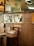 The scene depicts the interior of a pizzeria with a tiled wall and stacks of pizza boxes on chairs. Through a window, a person is seen preparing food in a kitchen. Various posters and drawings are attached to the wall, and a clock is visible above the window.