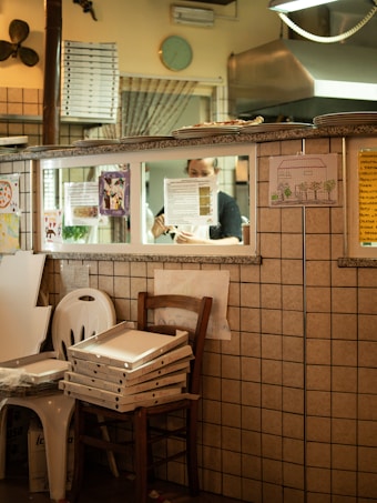 The scene depicts the interior of a pizzeria with a tiled wall and stacks of pizza boxes on chairs. Through a window, a person is seen preparing food in a kitchen. Various posters and drawings are attached to the wall, and a clock is visible above the window.