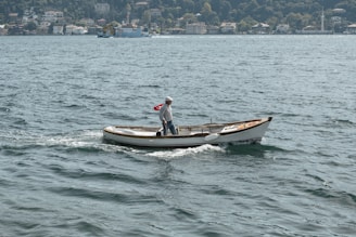 A small boat with a person on board is sailing across a body of water. The person is standing and looking towards the back of the boat. The boat features a Turkish flag at the stern. In the background, there is a view of a village or town with several houses and lush green trees.