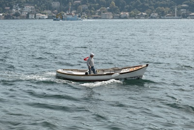 A small boat with a person on board is sailing across a body of water. The person is standing and looking towards the back of the boat. The boat features a Turkish flag at the stern. In the background, there is a view of a village or town with several houses and lush green trees.