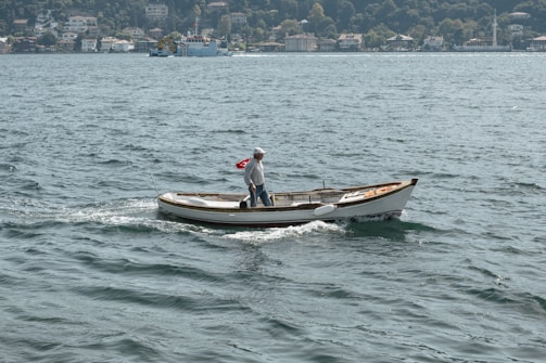 A small boat with a person on board is sailing across a body of water. The person is standing and looking towards the back of the boat. The boat features a Turkish flag at the stern. In the background, there is a view of a village or town with several houses and lush green trees.