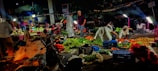 A bustling night market scene where vendors are selling a variety of vegetables. The atmosphere is lively with numerous baskets and crates filled with fresh produce like green leafy vegetables, tomatoes, and potatoes. Several people are engaged in buying and selling activities under bright lights, casting shadows around.