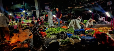A bustling night market scene where vendors are selling a variety of vegetables. The atmosphere is lively with numerous baskets and crates filled with fresh produce like green leafy vegetables, tomatoes, and potatoes. Several people are engaged in buying and selling activities under bright lights, casting shadows around.