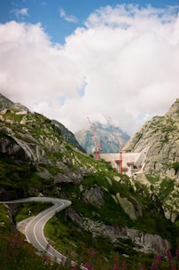 A winding road curves through a lush, mountainous landscape with steep rocky hillsides. In the background, red construction cranes rise above a dam structure set against the backdrop of towering peaks and a partially cloudy blue sky.