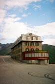 A large, older-looking stone building with green shutters and a metal roof sits at a curve in a mountain road. The building has signs indicating it is a hotel and restaurant. Surrounding the building are mountains and rolling hills under a partly cloudy sky.