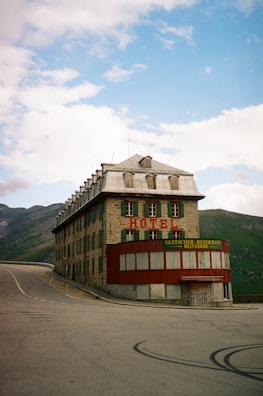 A large, older-looking stone building with green shutters and a metal roof sits at a curve in a mountain road. The building has signs indicating it is a hotel and restaurant. Surrounding the building are mountains and rolling hills under a partly cloudy sky.