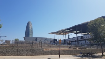 A modern urban landscape featuring a uniquely shaped, bullet-like skyscraper known as Torre Gl&ograve;ries against a clear blue sky. To the right, there is a market structure with a wavy, metallic roof and exposed beams. In the foreground, a paved sidewalk and a row of rental bicycles are visible.