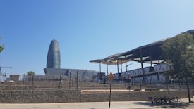 A modern urban landscape featuring a uniquely shaped, bullet-like skyscraper known as Torre Glòries against a clear blue sky. To the right, there is a market structure with a wavy, metallic roof and exposed beams. In the foreground, a paved sidewalk and a row of rental bicycles are visible.