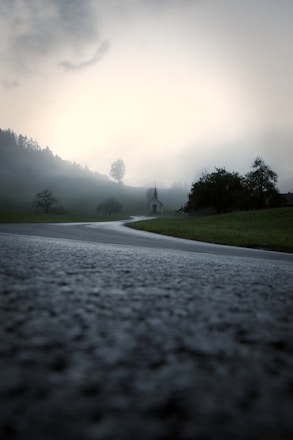 A winding road leads through a misty landscape with a small chapel in the distance under a luminous, overcast sky. Trees dot the scene, adding to the serene and mysterious atmosphere.