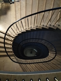 An overhead view of a spiral staircase with a central void, showcasing its curved form and metal railings. The steps are made of a light stone material, creating a dramatic pattern as they descend.