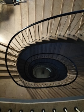 An overhead view of a spiral staircase with a central void, showcasing its curved form and metal railings. The steps are made of a light stone material, creating a dramatic pattern as they descend.