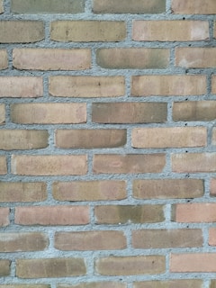 A close-up view of a brick wall composed of uniform rectangular bricks arranged in a horizontal pattern. The bricks are light brown and feature a rough texture, with grey mortar filling the gaps between them.