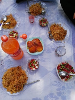 A picnic setup with multiple plates of fried noodles, small bowls of colorful salad, and a plate of orange-colored fried snacks. There is a large bottle of orange soda and cups placed on a patterned tablecloth.