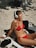 A woman in a red bikini is sitting on a pebble beach, wearing white headphones and basking in the sun with her eyes closed. Various items are placed around her, including a black cap, towels, and a bottle of sunscreen.