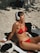 A woman in a red bikini is sitting on a pebble beach, wearing white headphones and basking in the sun with her eyes closed. Various items are placed around her, including a black cap, towels, and a bottle of sunscreen.