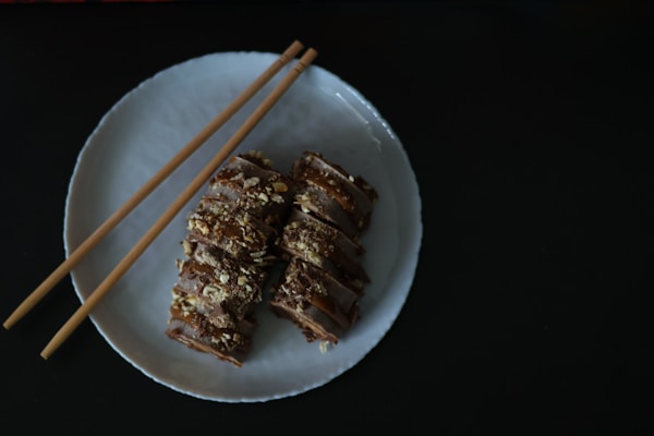 A white plate holds several pieces of sliced sushi garnished with crumbs, placed elegantly alongside a pair of wooden chopsticks. The dark background enhances the presentation of the food.