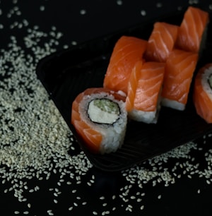 A black tray contains slices of sushi rolls topped with fresh salmon. The rolls include ingredients like rice, cream cheese, and cucumber, and are set against a dark background sprinkled with sesame seeds.