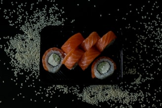 A black tray is filled with neatly arranged sushi rolls topped with fresh orange salmon slices. The rolls contain rice, avocado, and cream cheese, wrapped in seaweed. Scattered around the tray are white sesame seeds on a dark background.