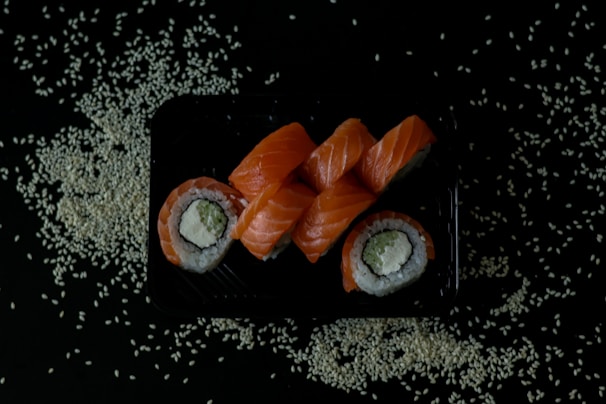 A black tray is filled with neatly arranged sushi rolls topped with fresh orange salmon slices. The rolls contain rice, avocado, and cream cheese, wrapped in seaweed. Scattered around the tray are white sesame seeds on a dark background.