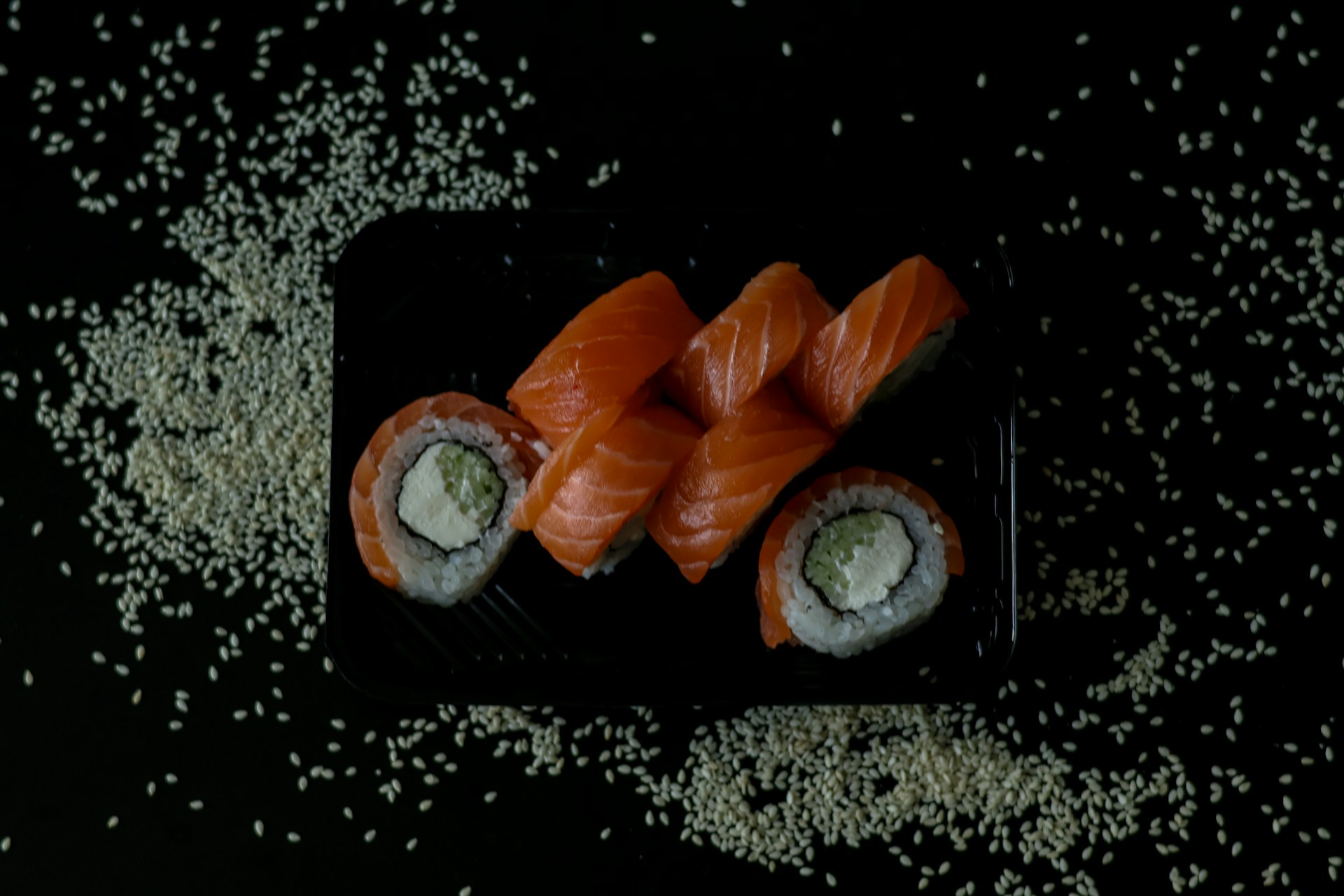 A black tray is filled with neatly arranged sushi rolls topped with fresh orange salmon slices. The rolls contain rice, avocado, and cream cheese, wrapped in seaweed. Scattered around the tray are white sesame seeds on a dark background.