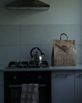 A modern kitchen stove with a kettle on one of the burners, a brown paper bag with the logo 'Dart Sushi', and a towel hanging on the oven door. The stovetop has a minimalist design, and the kitchen has an extractor hood above the stove.