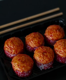 Six pieces of sushi are arranged in a black tray, topped with what appears to be a roasted or spicy sauce. The sushi pieces have a reddish-orange topping, possibly made from fish roe or a spicy mix, atop a layer of rice.