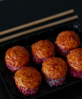 Six pieces of sushi are arranged in a black tray, topped with what appears to be a roasted or spicy sauce. The sushi pieces have a reddish-orange topping, possibly made from fish roe or a spicy mix, atop a layer of rice.