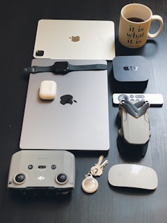 A collection of electronic devices and accessories are neatly arranged on a dark surface. Items include a tablet, a smartwatch, wireless earbuds, a TV box with remote, a drone, a controller, a set of keys with a key ring, a wireless mouse, and a coffee mug with text.
