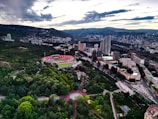 Aerial view of a city with a large, circular stadium surrounded by greenery and modern buildings. The landscape features rolling hills in the background under a partly cloudy sky, and numerous tall buildings are scattered throughout the city. Roads weave through the urban and green spaces, creating a striking contrast between nature and urban development.