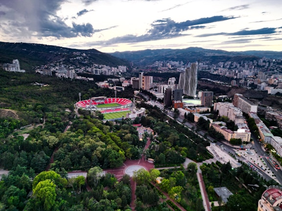 Aerial view of a city with a large, circular stadium surrounded by greenery and modern buildings. The landscape features rolling hills in the background under a partly cloudy sky, and numerous tall buildings are scattered throughout the city. Roads weave through the urban and green spaces, creating a striking contrast between nature and urban development.