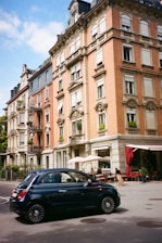 A classic European street scene features a red-brick building with ornate architectural details. The building has several floors with white window frames and decorative molding. A small cafe with outdoor seating occupies the ground level, with people seated under beige umbrellas. In the foreground, a dark blue compact car drives by on the street.