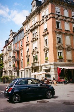 A classic European street scene features a red-brick building with ornate architectural details. The building has several floors with white window frames and decorative molding. A small cafe with outdoor seating occupies the ground level, with people seated under beige umbrellas. In the foreground, a dark blue compact car drives by on the street.