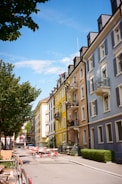 A street with colorful residential buildings lining both sides. The buildings are painted in shades of blue, yellow, and beige, with balconies and decorative window shutters. Trees with green foliage are along the street, and there are construction barriers and materials present on the road.