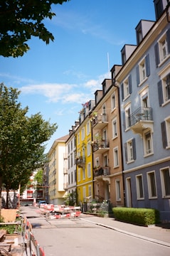 A street with colorful residential buildings lining both sides. The buildings are painted in shades of blue, yellow, and beige, with balconies and decorative window shutters. Trees with green foliage are along the street, and there are construction barriers and materials present on the road.