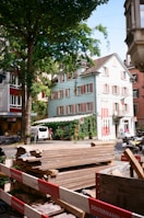 A street scene featuring a construction site with wooden planks piled behind a barrier marked with the name of a construction company. In the background, there is a light blue building with red and white shutters, partly covered in ivy. Other buildings and a van are also visible, along with trees and bicycles parked nearby.