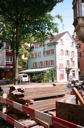 A street scene featuring a construction site with wooden planks piled behind a barrier marked with the name of a construction company. In the background, there is a light blue building with red and white shutters, partly covered in ivy. Other buildings and a van are also visible, along with trees and bicycles parked nearby.