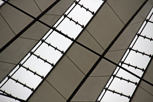 A close-up view of an architectural structure featuring symmetrical, triangular panels made of light fabric with supporting black metal beams. The panels and beams create a geometric pattern against a cloudy sky.