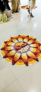 A colorful flower arrangement in a circular pattern, displayed on a polished floor. People in traditional attire are visible, partially captured, suggesting a cultural or festive setting. The flower design includes concentric circles and petal shapes using red, yellow, orange, and white petals.