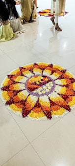 A colorful flower arrangement in a circular pattern, displayed on a polished floor. People in traditional attire are visible, partially captured, suggesting a cultural or festive setting. The flower design includes concentric circles and petal shapes using red, yellow, orange, and white petals.