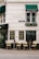 A quaint street-side restaurant or cafe featuring outdoor seating with wooden tables and rattan chairs arranged neatly. The establishment’s exterior has white walls with green awnings. Signage on the walls advertises various spirits, wines, beers, ales, and stouts. Plant arrangements add a touch of greenery.