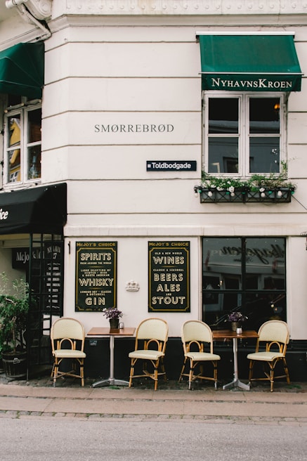 A quaint street-side restaurant or cafe featuring outdoor seating with wooden tables and rattan chairs arranged neatly. The establishment’s exterior has white walls with green awnings. Signage on the walls advertises various spirits, wines, beers, ales, and stouts. Plant arrangements add a touch of greenery.