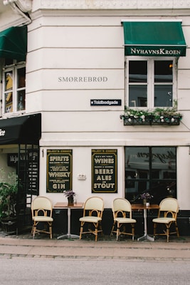 A quaint street-side restaurant or cafe featuring outdoor seating with wooden tables and rattan chairs arranged neatly. The establishment’s exterior has white walls with green awnings. Signage on the walls advertises various spirits, wines, beers, ales, and stouts. Plant arrangements add a touch of greenery.